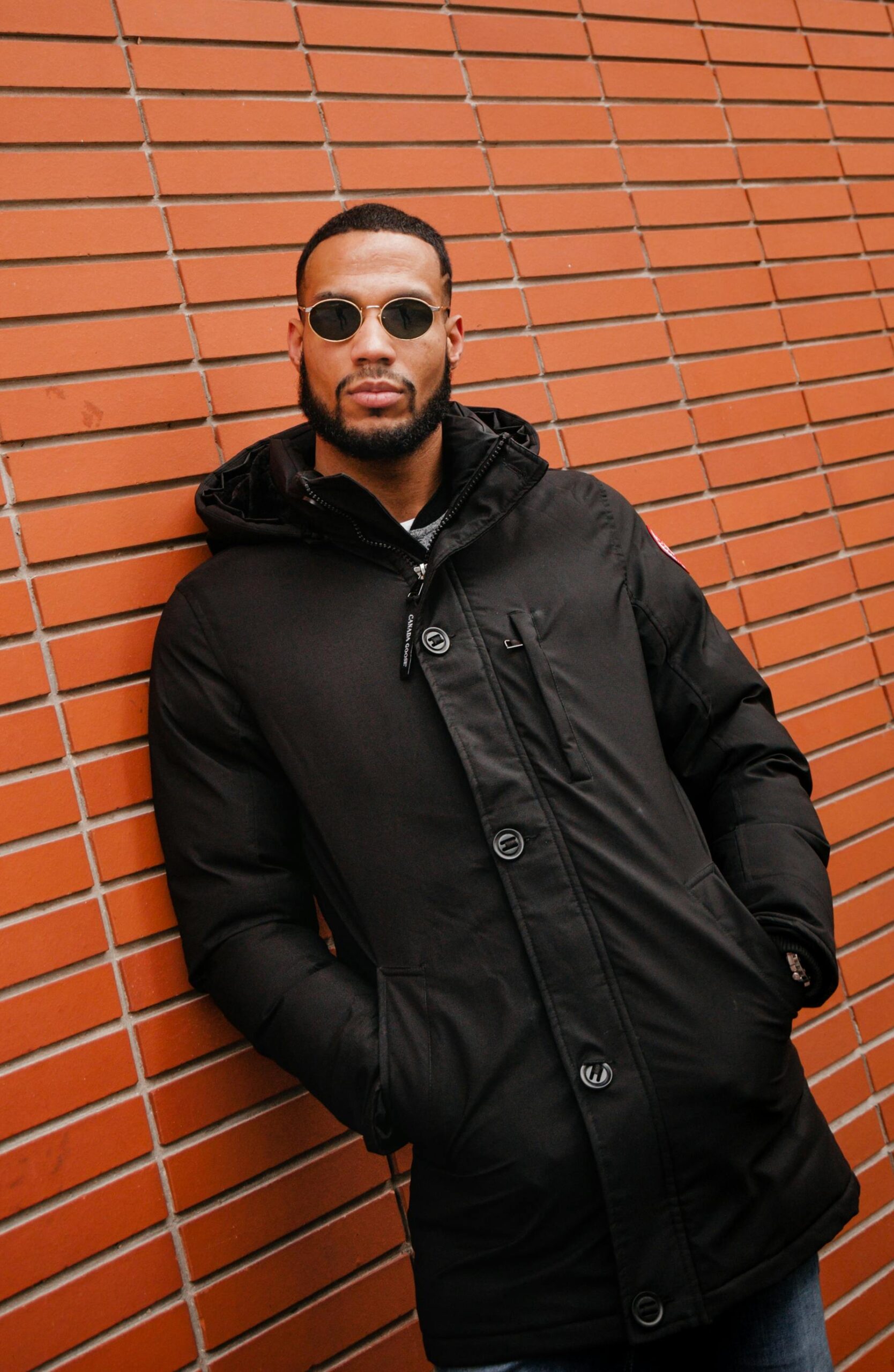 Stylish man wearing sunglasses and black coat leans against a brick wall, embodying urban fashion.